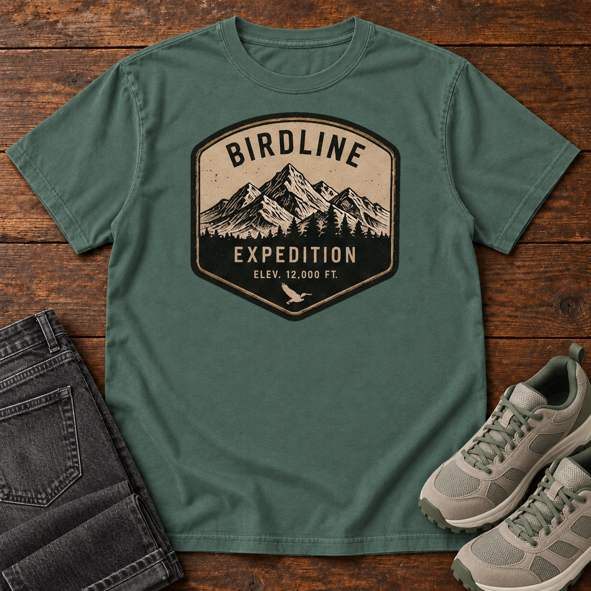 Birdline Expedition T-Shirt in Blue Spruce, front laid flat on wood, featuring a tan-and-black expedition badge with mountain peaks, pine trees and BIRDLINE lettering.