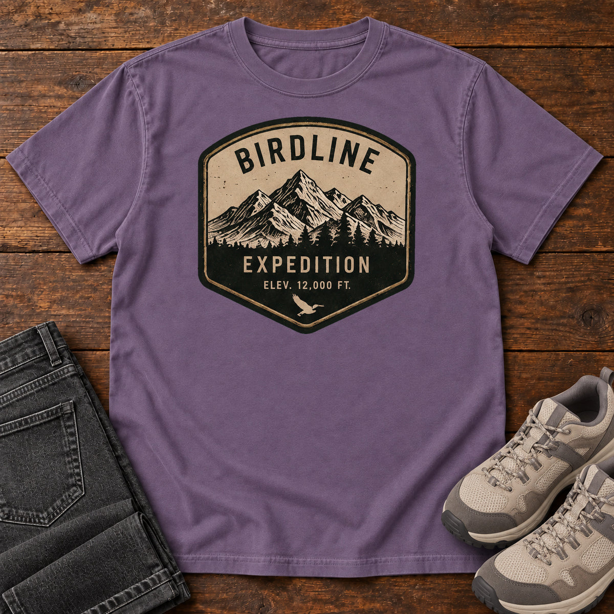 Birdline Expedition T-Shirt in Grape, front laid flat on wood, featuring a tan-and-black expedition badge with mountain peaks, pine trees and BIRDLINE lettering.