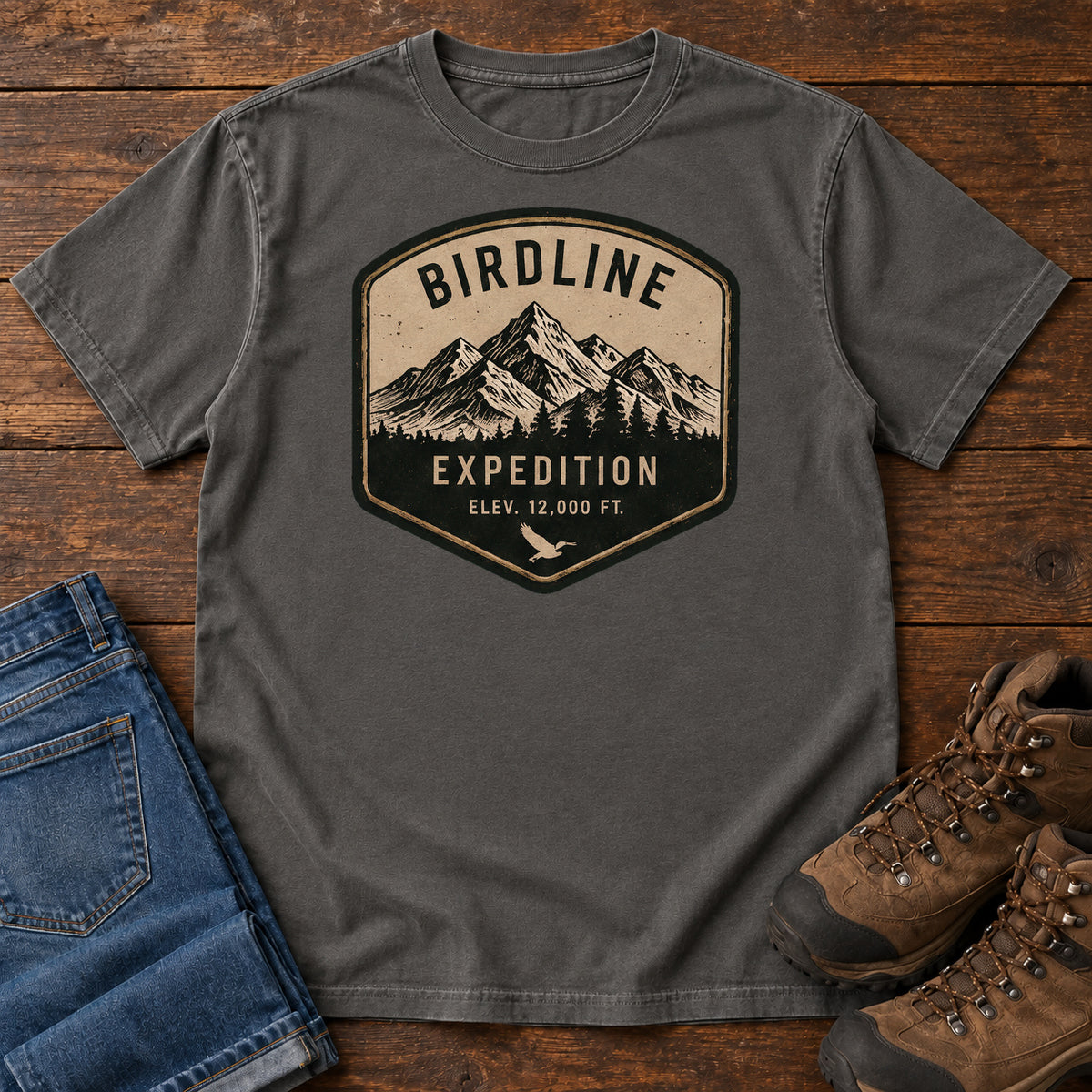 Birdline Expedition T-Shirt in Pepper, front laid flat on wood, featuring a tan-and-black expedition badge with mountain peaks, pine trees and BIRDLINE lettering.