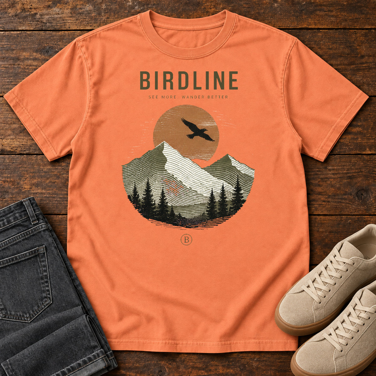 Terracotta Wander Better T-Shirt with a soaring bird, orange sun and layered mountain-and-forest graphic, front flat lay on wood.