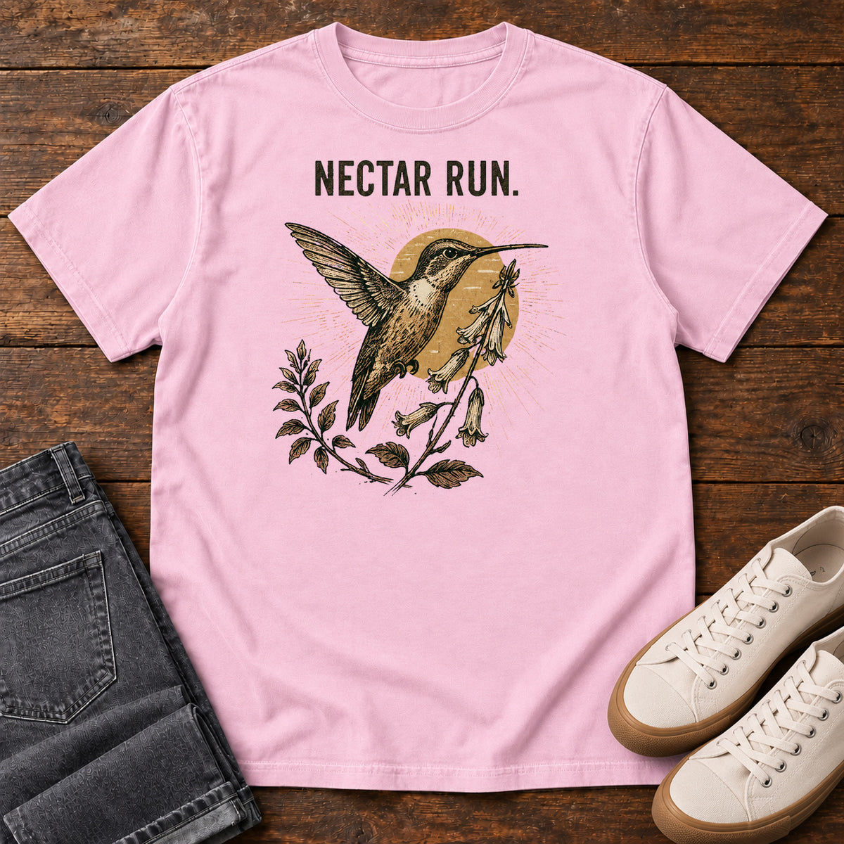 Blossom Nectar Run Sunburst T-Shirt with a long-beaked bird hovering beside flowers against a golden sunburst; front flat lay on wood.