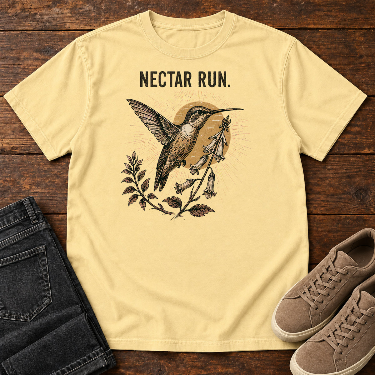 Butter Nectar Run Sunburst T-Shirt with a long-beaked bird hovering beside flowers against a golden sunburst; front flat lay on wood.