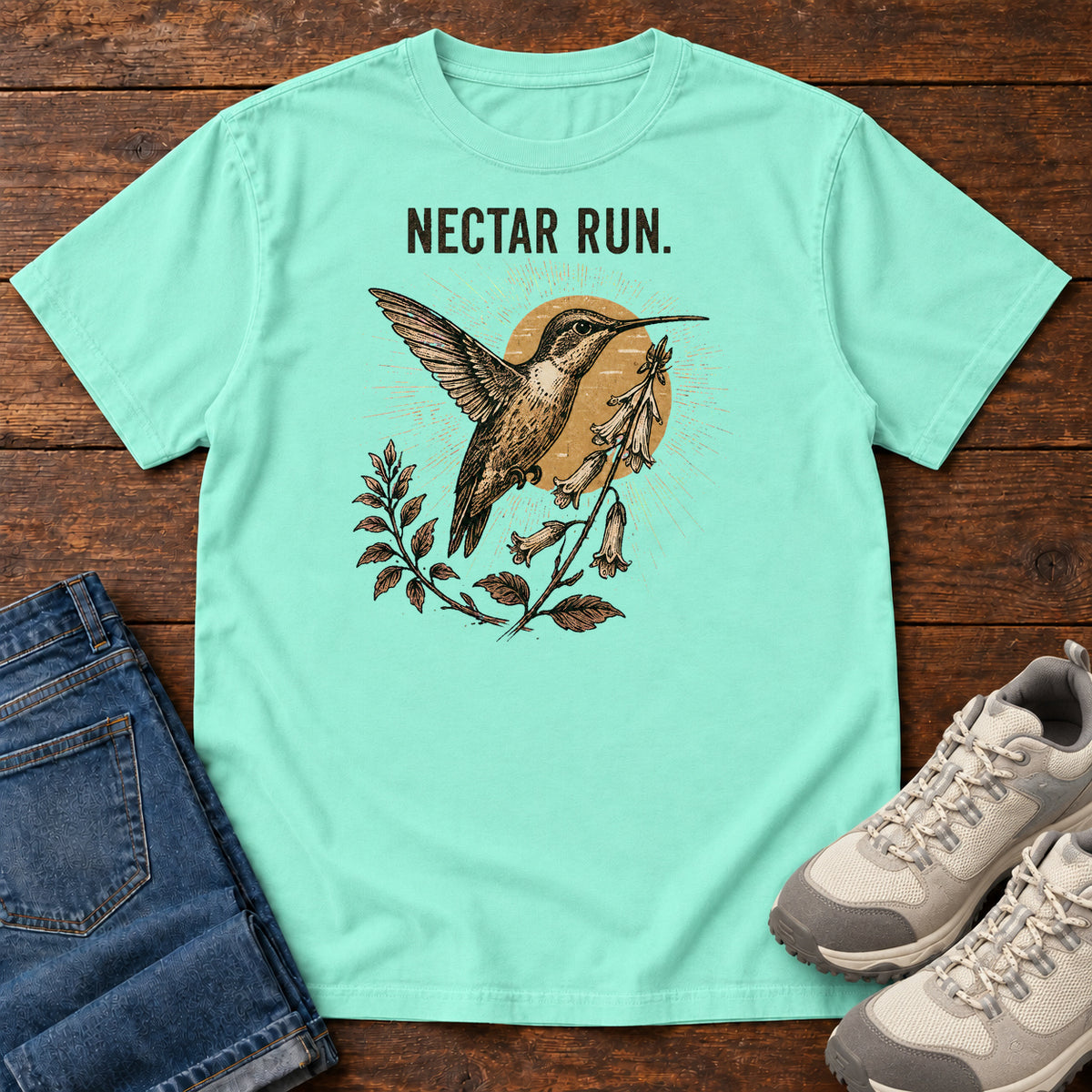 Chalky Mint Nectar Run Sunburst T-Shirt with a long-beaked bird hovering beside flowers against a golden sunburst; front flat lay on wood.