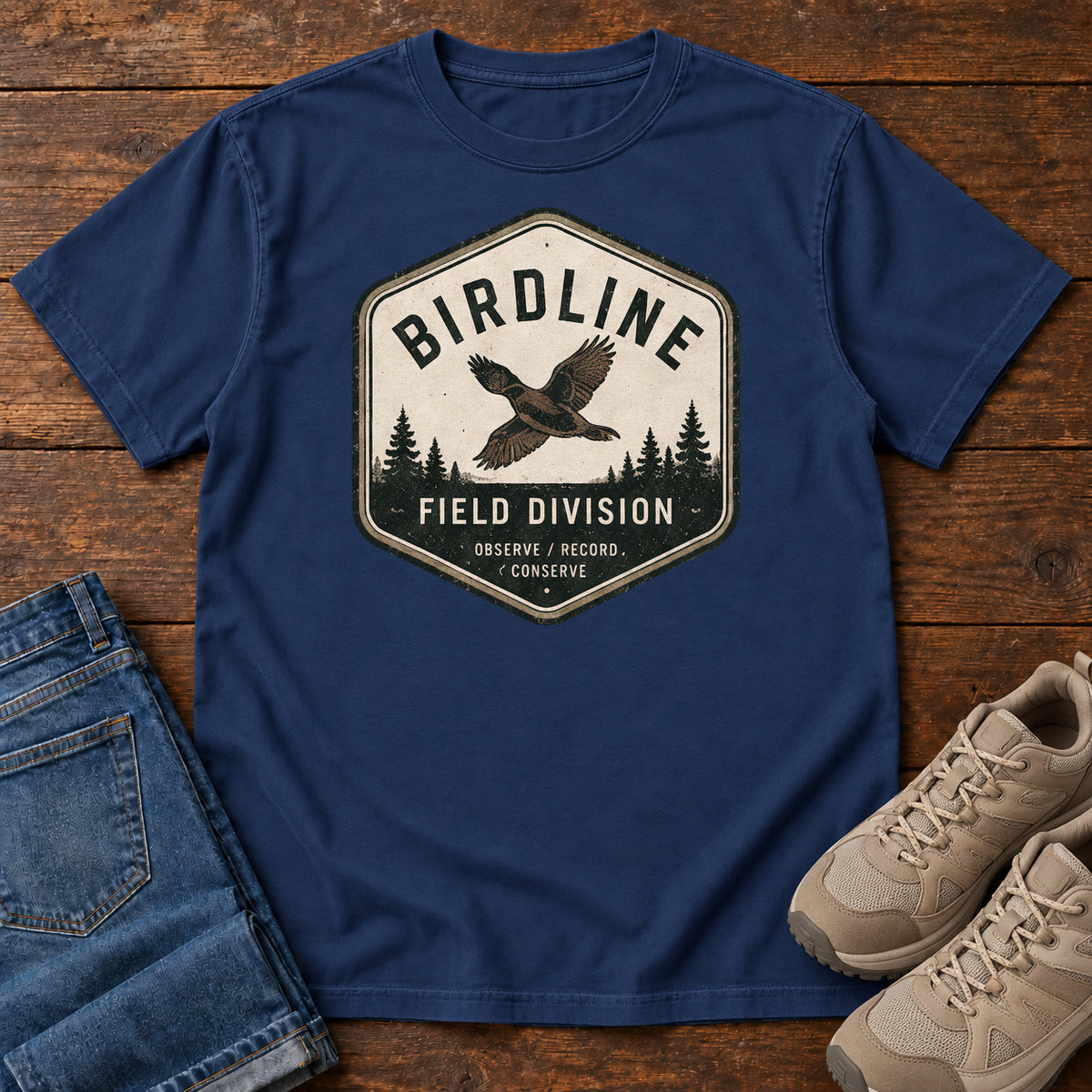 True Navy Birdline Field Division T-Shirt with a cream badge showing a flying bird over pine trees; front flat lay on wood.