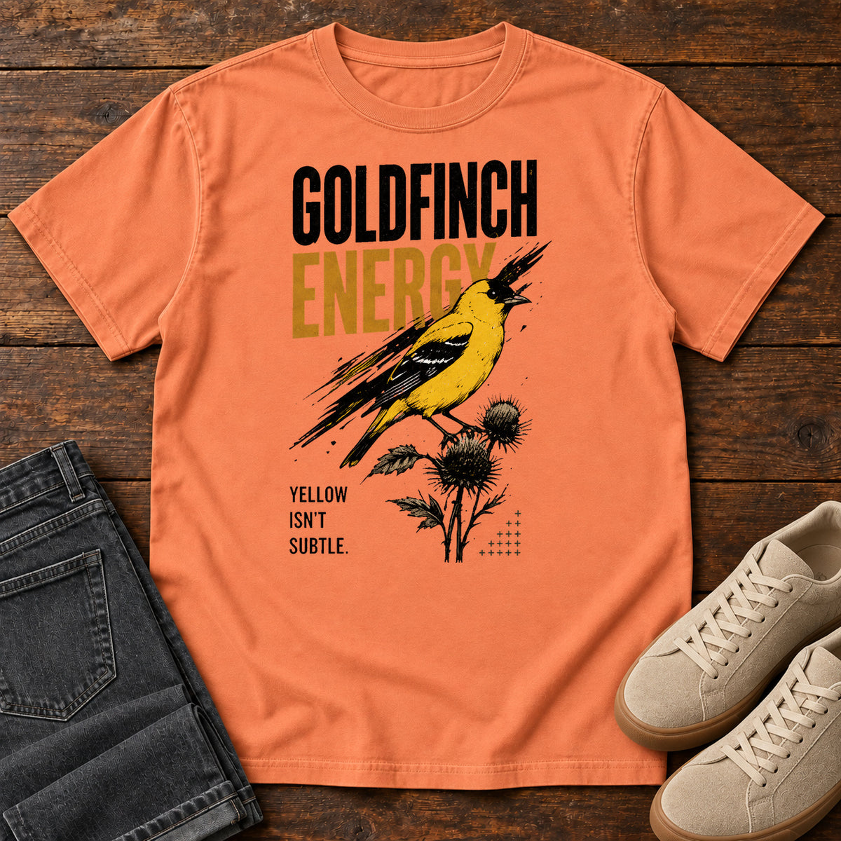 Terracotta Goldfinch Energy T-shirt with a yellow bird on thistles and Goldfinch Energy lettering, laid flat on a wooden surface.