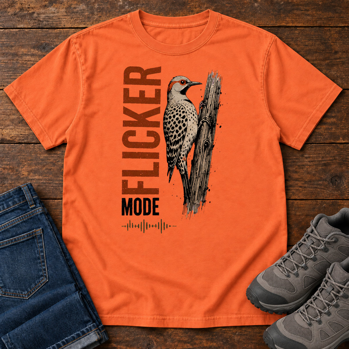 Burnt Orange Flicker Mode T-shirt with a spotted bird on a tree trunk and Flicker Mode lettering, laid flat on a wooden surface.