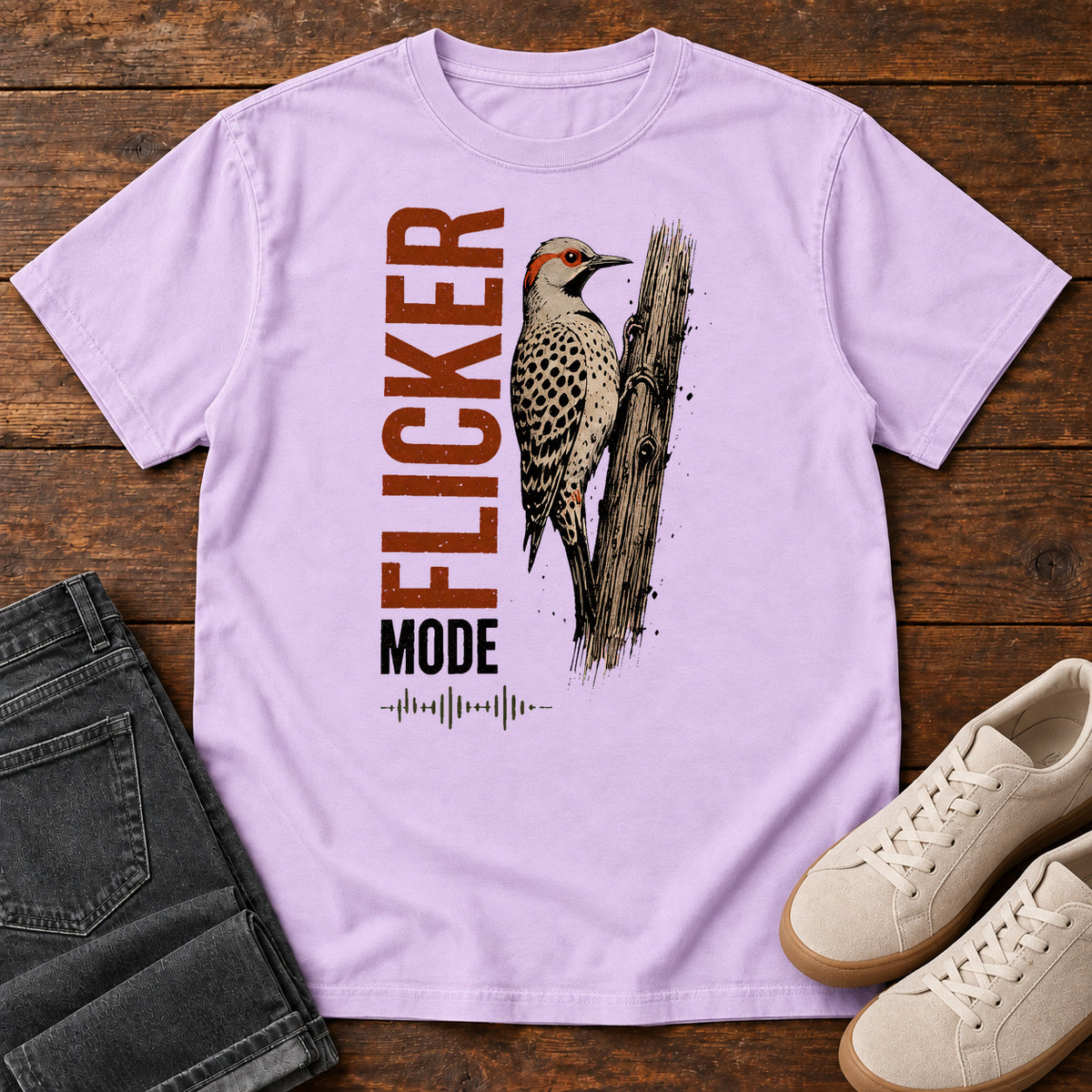 Orchid Flicker Mode T-shirt with a spotted bird on a tree trunk and Flicker Mode lettering, laid flat on a wooden surface.