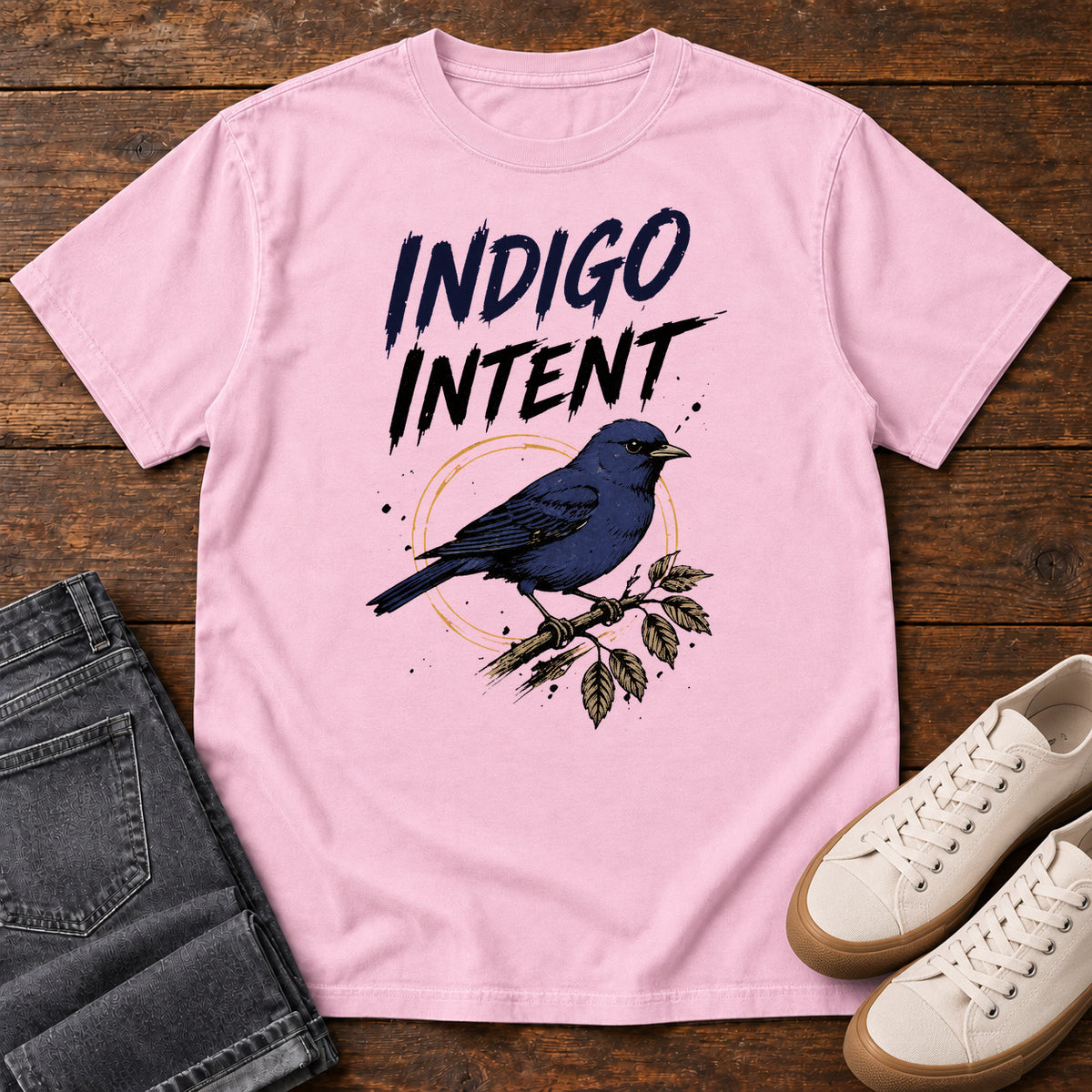 Blossom Indigo Intent T-shirt with a blue bird on a branch and Indigo Intent lettering, laid flat on a wooden surface.