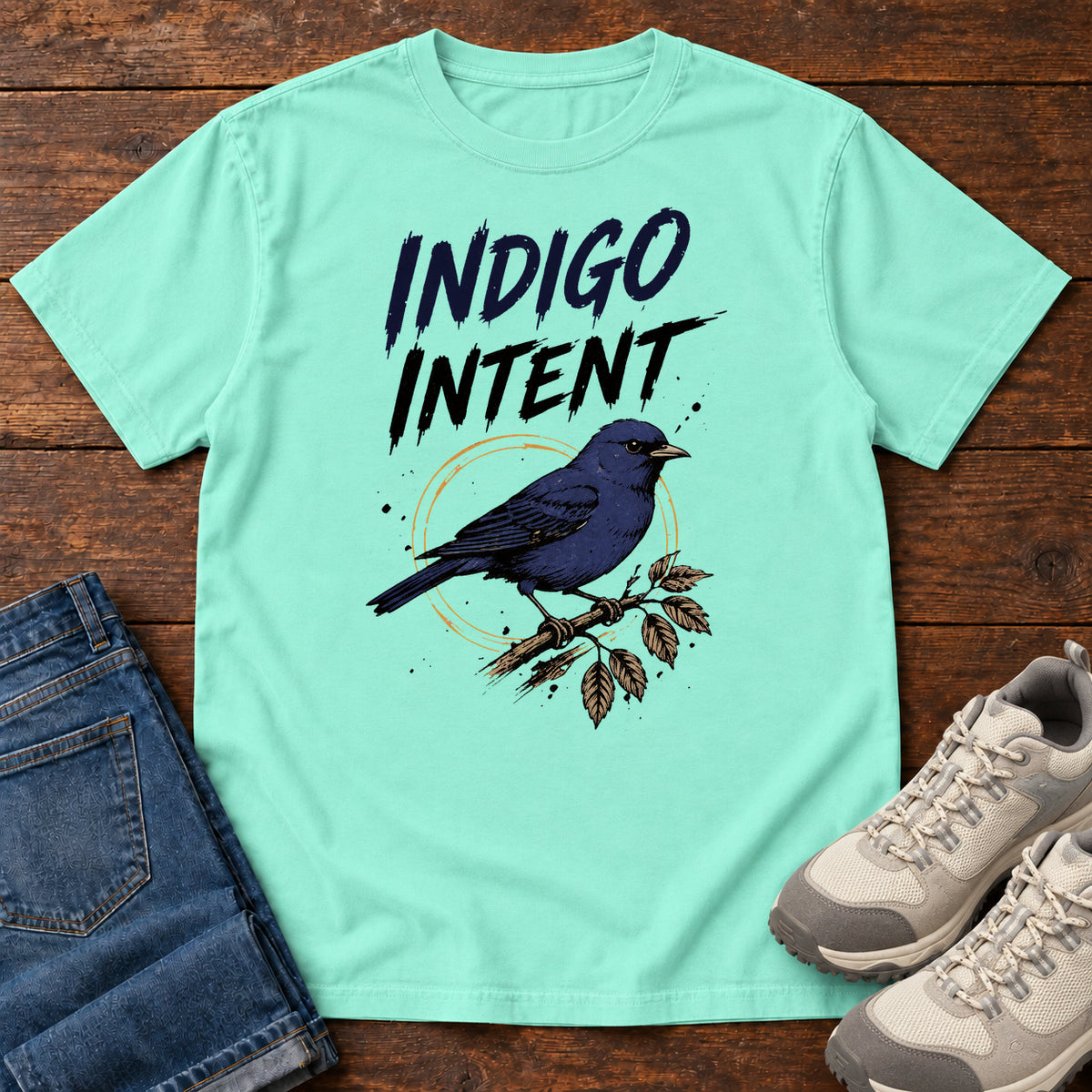 Chalky Mint Indigo Intent T-shirt with a blue bird on a branch and Indigo Intent lettering, laid flat on a wooden surface.