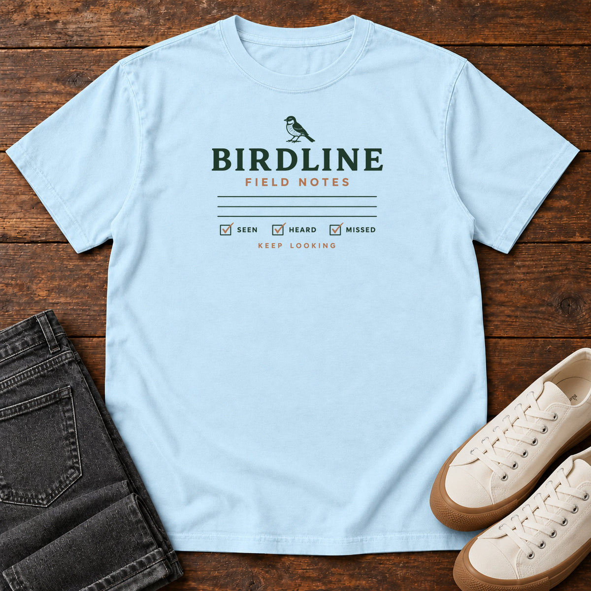 Chambray Field Notes T-shirt with a small bird above Birdline Field Notes lettering, ruled lines and checkboxes, front flat lay on wood.