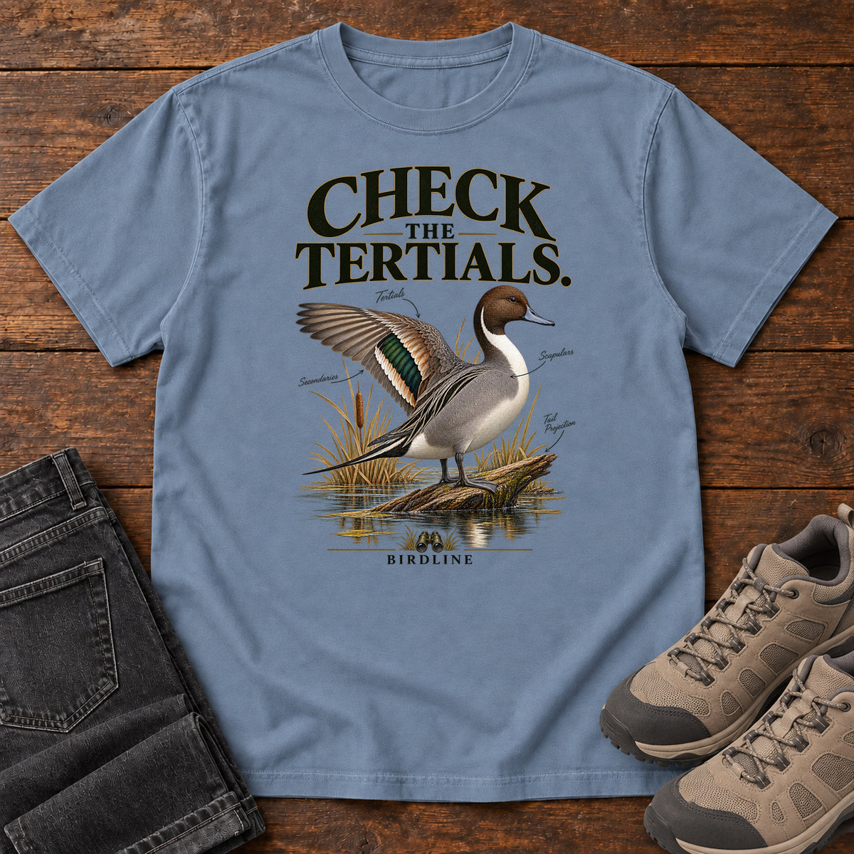 Blue Jean Check the Tertials Duck T-Shirt with a duck raising one wing beside reeds and water; front flat lay on wood.