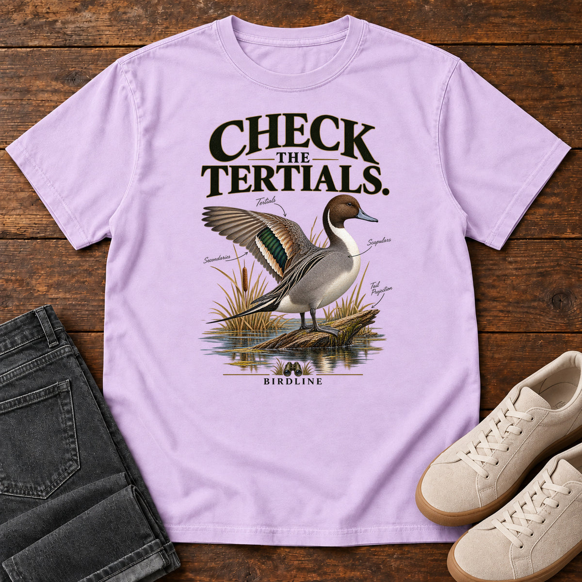 Orchid Check the Tertials Duck T-Shirt with a duck raising one wing beside reeds and water; front flat lay on wood.