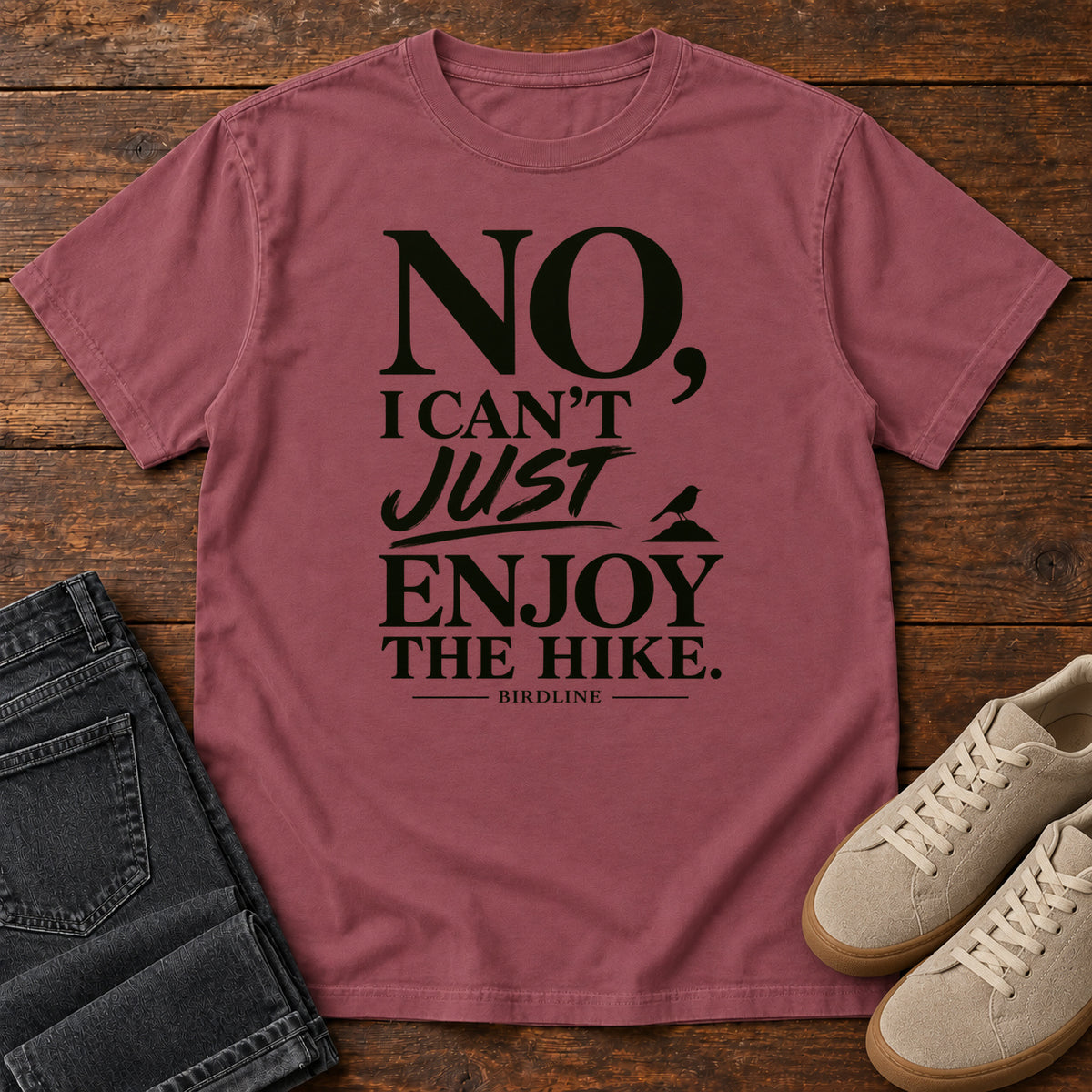 Brick No I Can't Just Enjoy the Hike T-Shirt with bold black lettering and a small bird silhouette, front flat lay on wood.