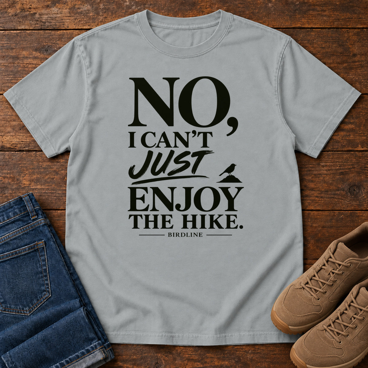Granite No I Can't Just Enjoy the Hike T-Shirt with bold black lettering and a small bird silhouette, front flat lay on wood.