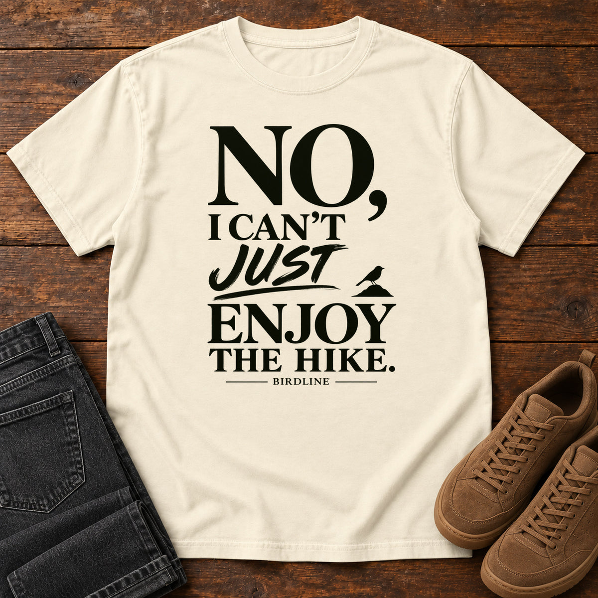 Ivory No I Can't Just Enjoy the Hike T-Shirt with bold black lettering and a small bird silhouette, front flat lay on wood.