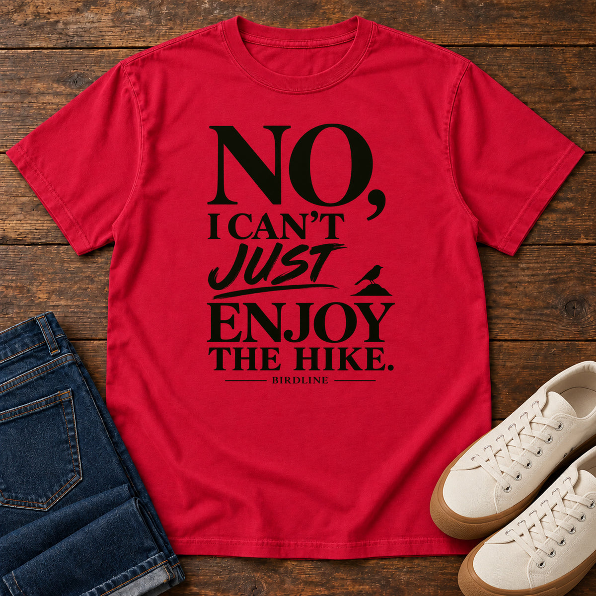 Red No I Can't Just Enjoy the Hike T-Shirt with bold black lettering and a small bird silhouette, front flat lay on wood.
