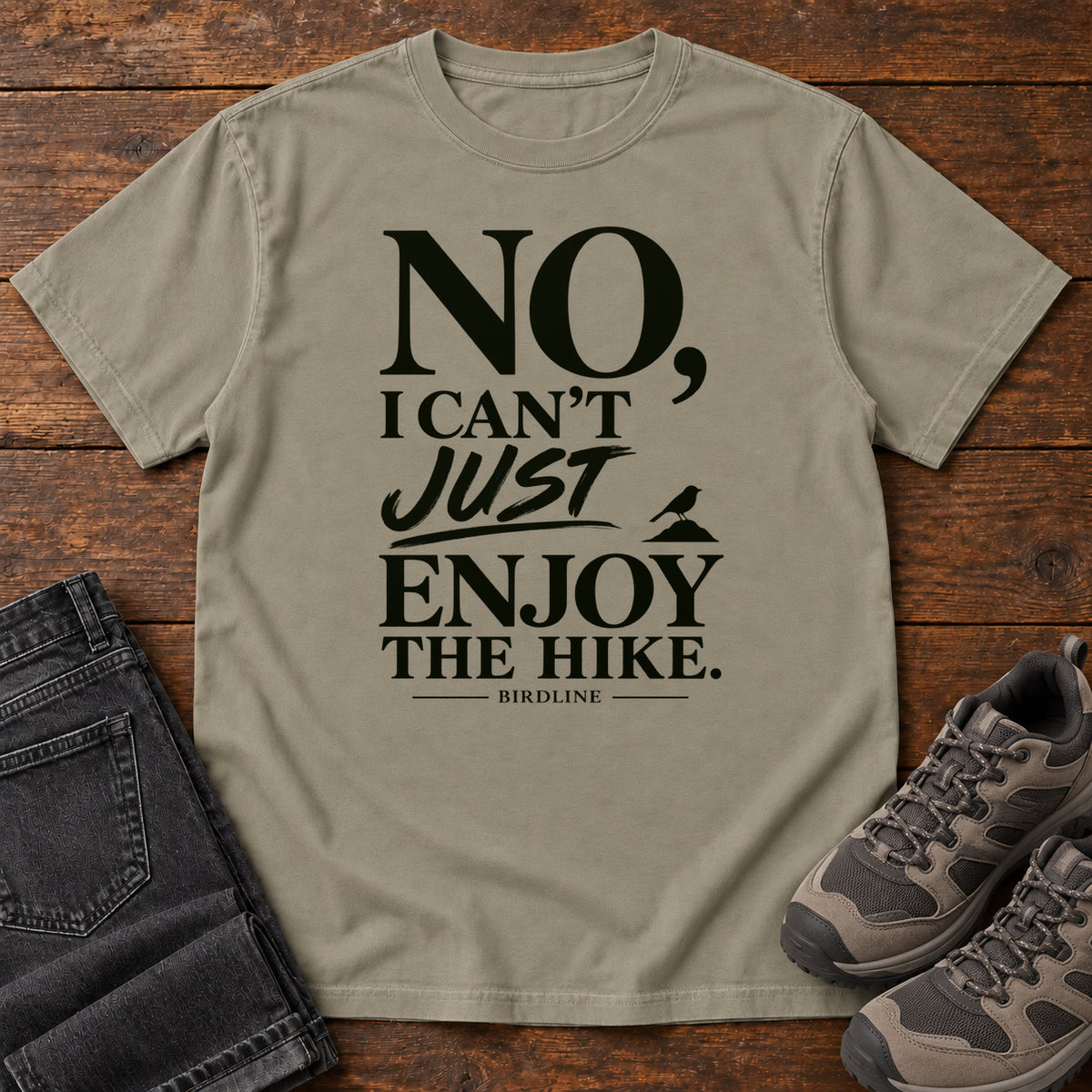 Sandstone No I Can't Just Enjoy the Hike T-Shirt with bold black lettering and a small bird silhouette, front flat lay on wood.