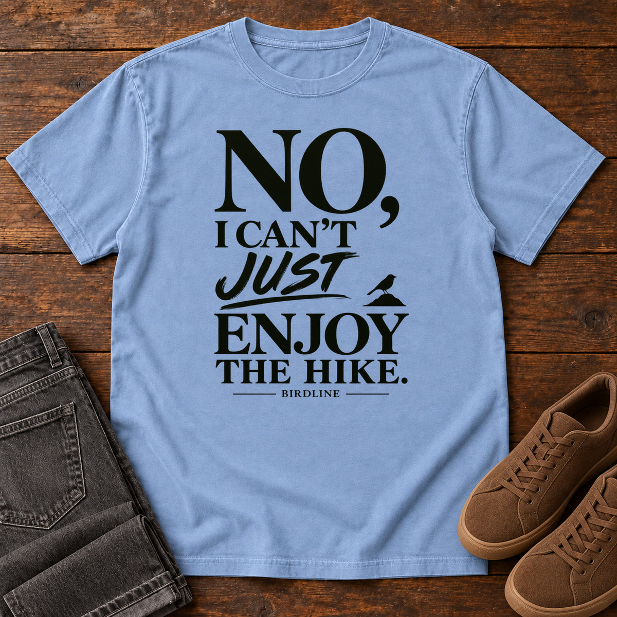 Washed Denim No I Can't Just Enjoy the Hike T-Shirt with bold black lettering and a small bird silhouette, front flat lay on wood.