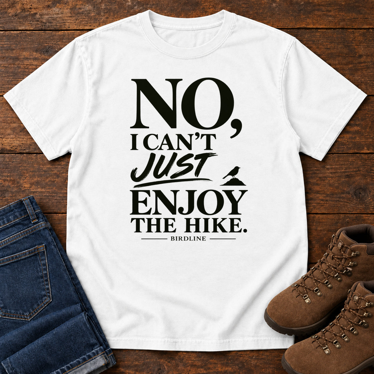 White No I Can't Just Enjoy the Hike T-Shirt with bold black lettering and a small bird silhouette, front flat lay on wood.
