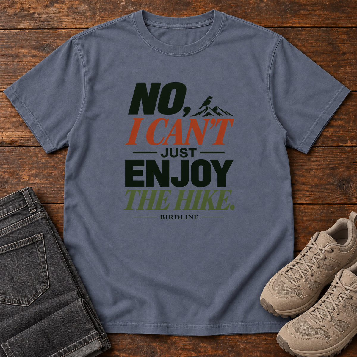 No I Can't Just Enjoy the Hike T-Shirt – Color Block in Denim, front laid flat on wood, featuring stacked green and rust slogan lettering and a small mountain graphic.