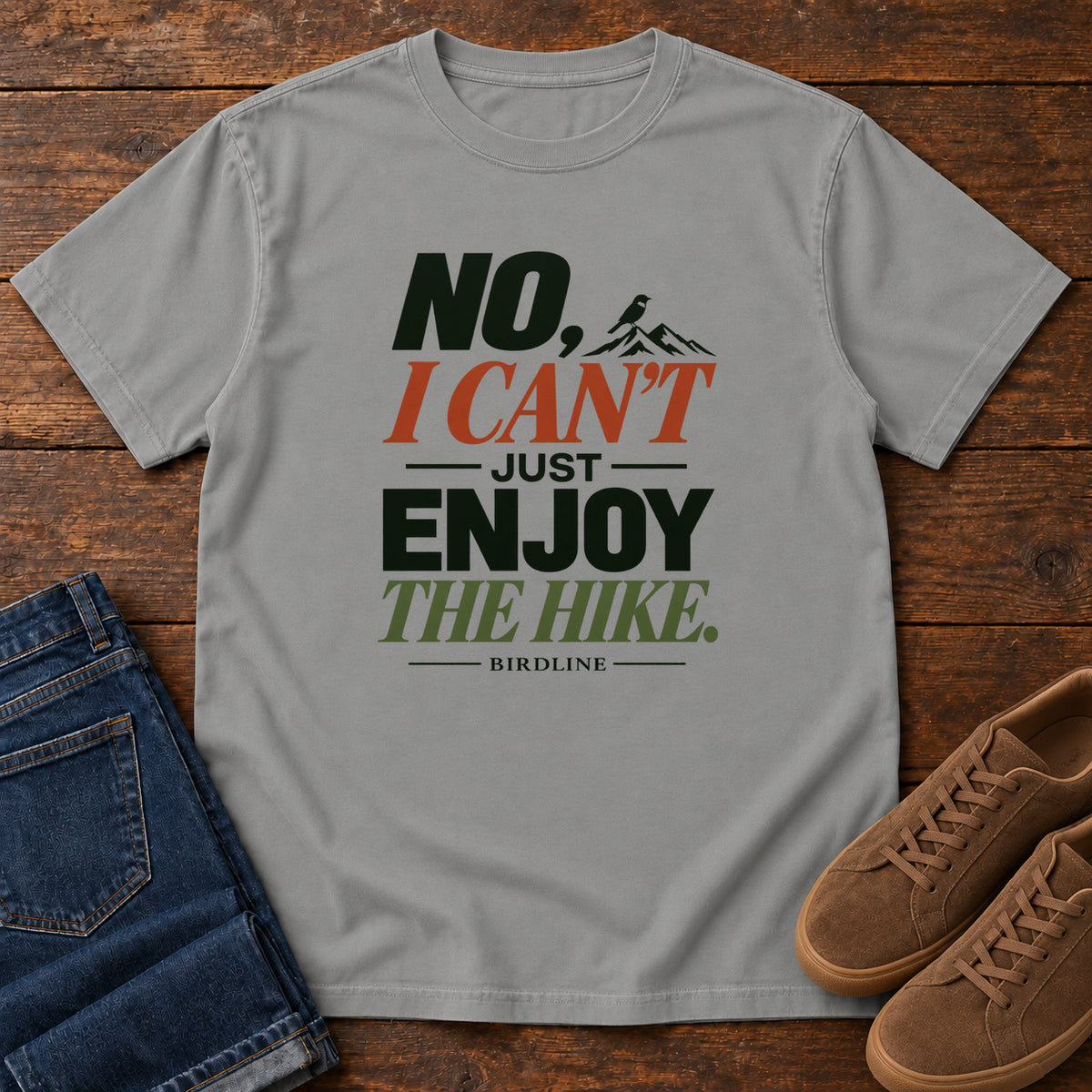 No I Can't Just Enjoy the Hike T-Shirt – Color Block in Grey, front laid flat on wood, featuring stacked green and rust slogan lettering and a small mountain graphic.