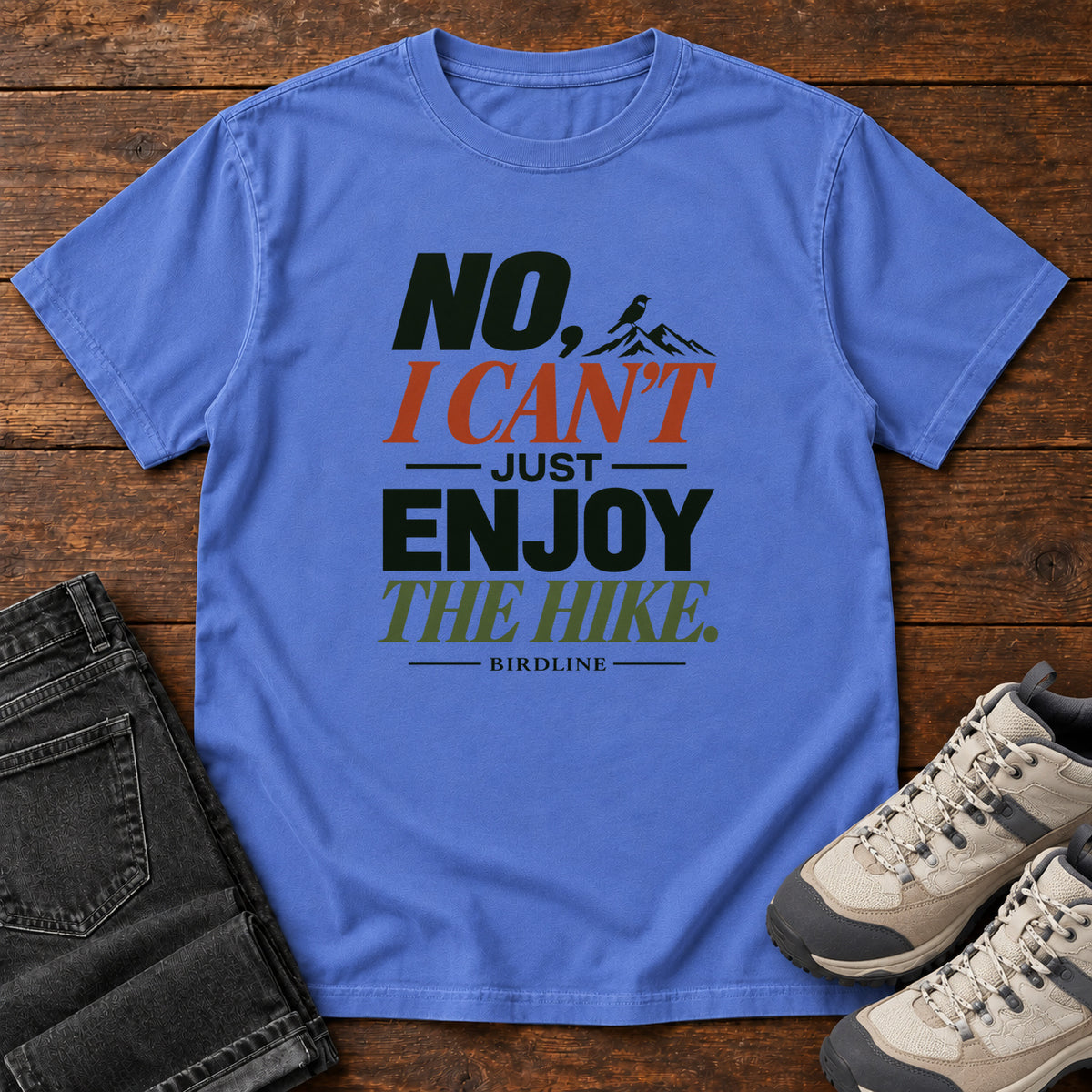 No I Can't Just Enjoy the Hike T-Shirt – Color Block in Mystic Blue, front laid flat on wood, featuring stacked green and rust slogan lettering and a small mountain graphic.