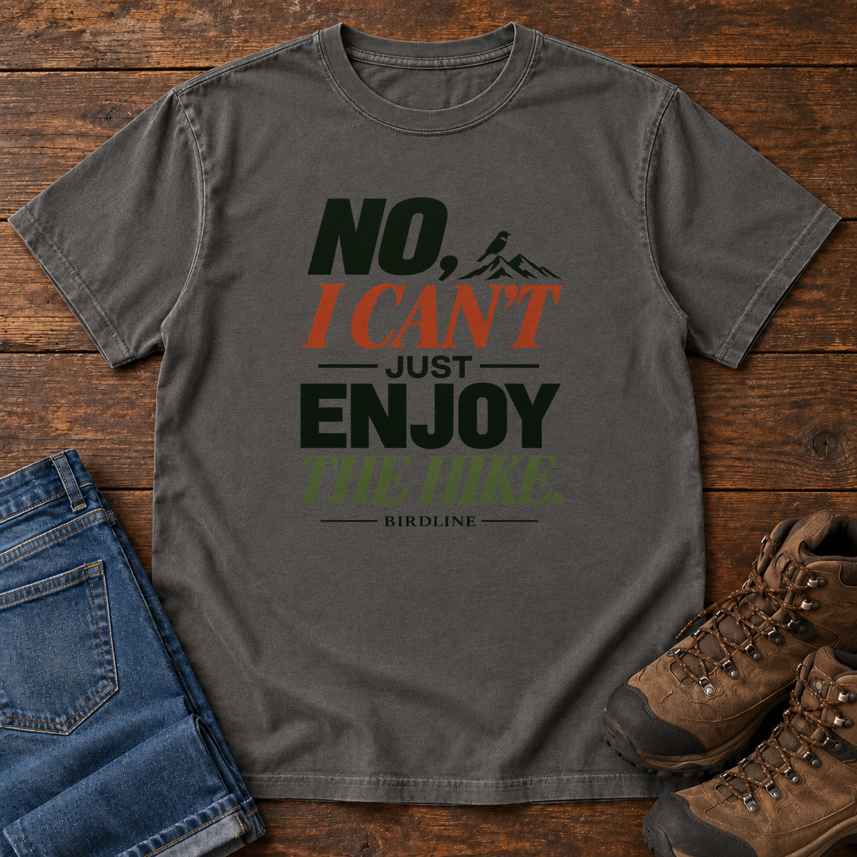 No I Can't Just Enjoy the Hike T-Shirt – Color Block in Pepper, front laid flat on wood, featuring stacked green and rust slogan lettering and a small mountain graphic.
