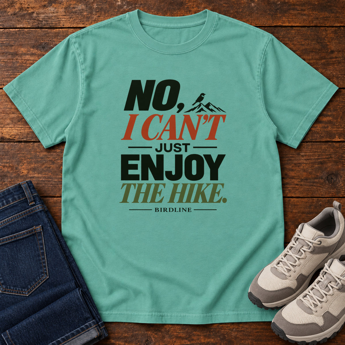 No I Can't Just Enjoy the Hike T-Shirt – Color Block in Seafoam, front laid flat on wood, featuring stacked green and rust slogan lettering and a small mountain graphic.