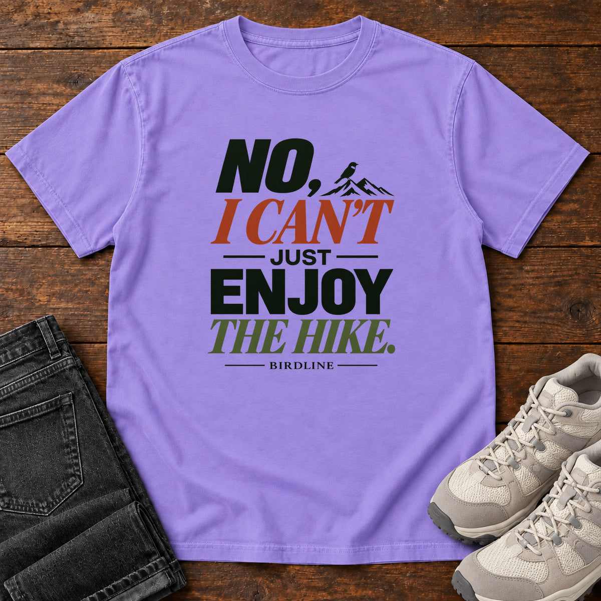 No I Can't Just Enjoy the Hike T-Shirt – Color Block in Violet, front laid flat on wood, featuring stacked green and rust slogan lettering and a small mountain graphic.