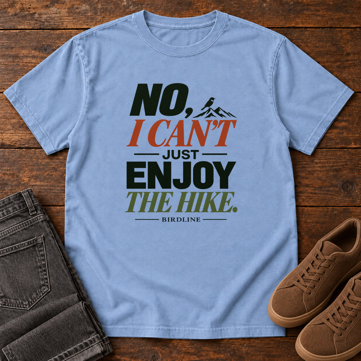 No I Can't Just Enjoy the Hike T-Shirt – Color Block in Washed Denim, front laid flat on wood, featuring stacked green and rust slogan lettering and a small mountain graphic.