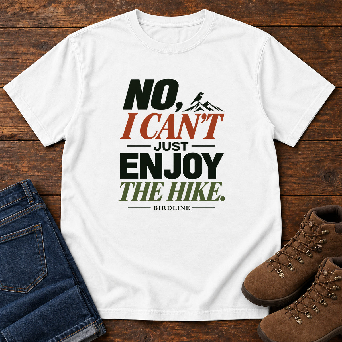No I Can't Just Enjoy the Hike T-Shirt – Color Block in White, front laid flat on wood, featuring stacked green and rust slogan lettering and a small mountain graphic.