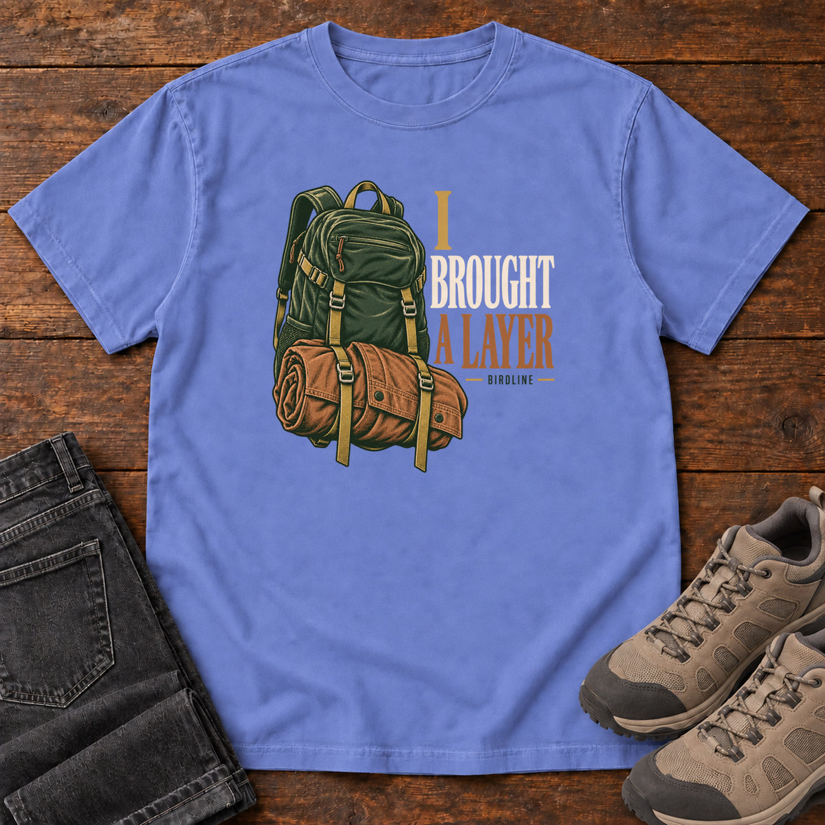 Flo Blue I Brought a Layer T-Shirt with a green hiking backpack with a brown roll strapped beneath it beside cream and gold lettering, front flat lay on wood.
