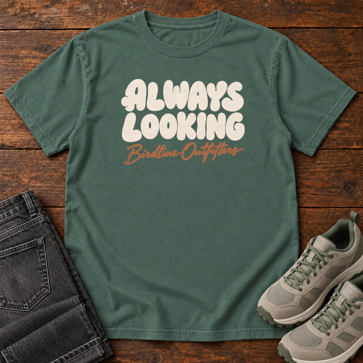 Blue Spruce Always Looking T-shirt with cream bubble lettering above an orange Birdline Outfitters script; front flat lay on wood.
