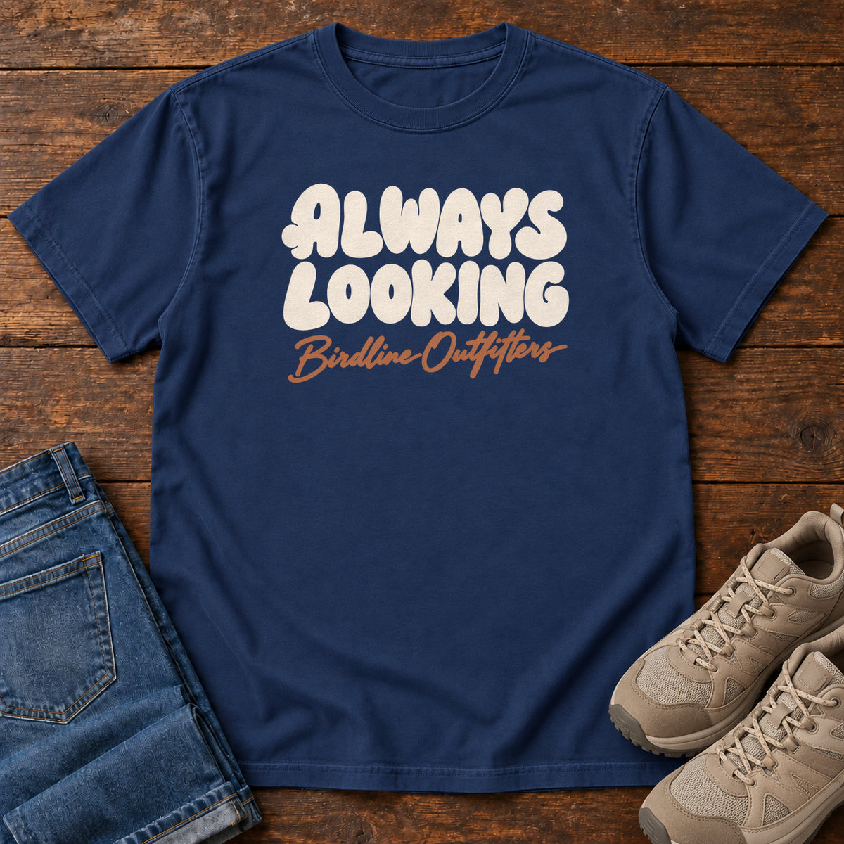 True Navy Always Looking T-shirt with cream bubble lettering above an orange Birdline Outfitters script; front flat lay on wood.