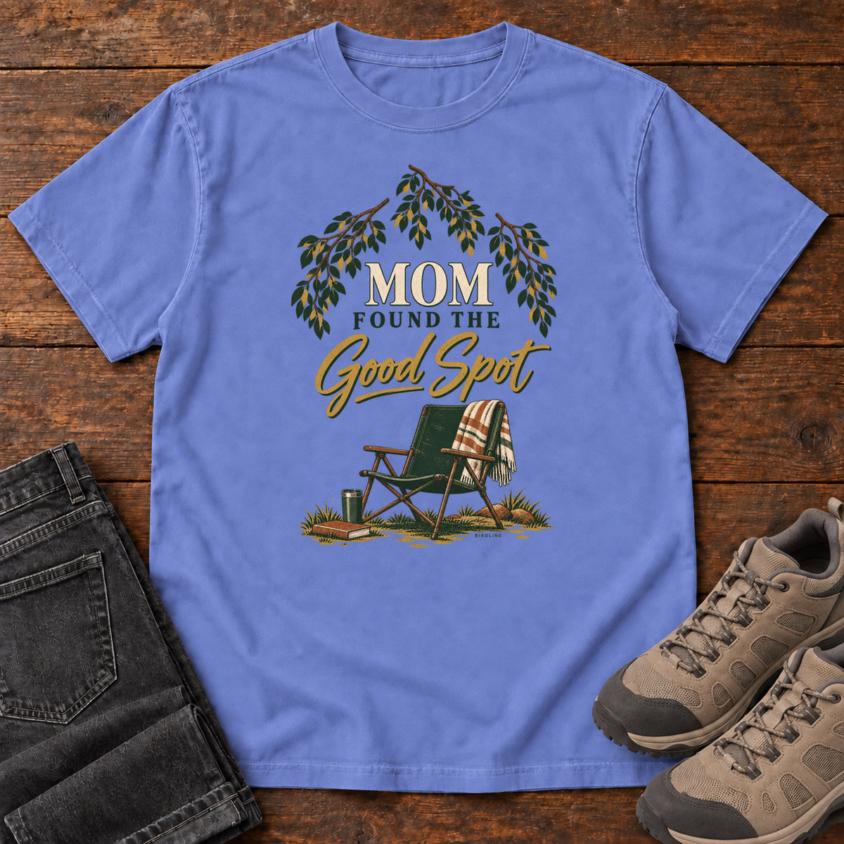 Flo Blue Mom Found the Good Spot T-Shirt with a folding camp chair, plaid blanket, leafy branches and gold script, front flat lay on wood.