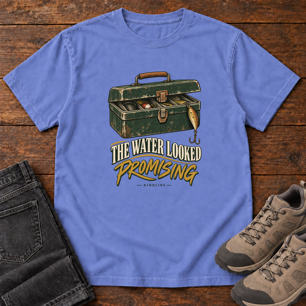 Flo Blue The Water Looked Promising T-Shirt with an open green tackle box and cream and gold slogan lettering; front flat lay on wood.