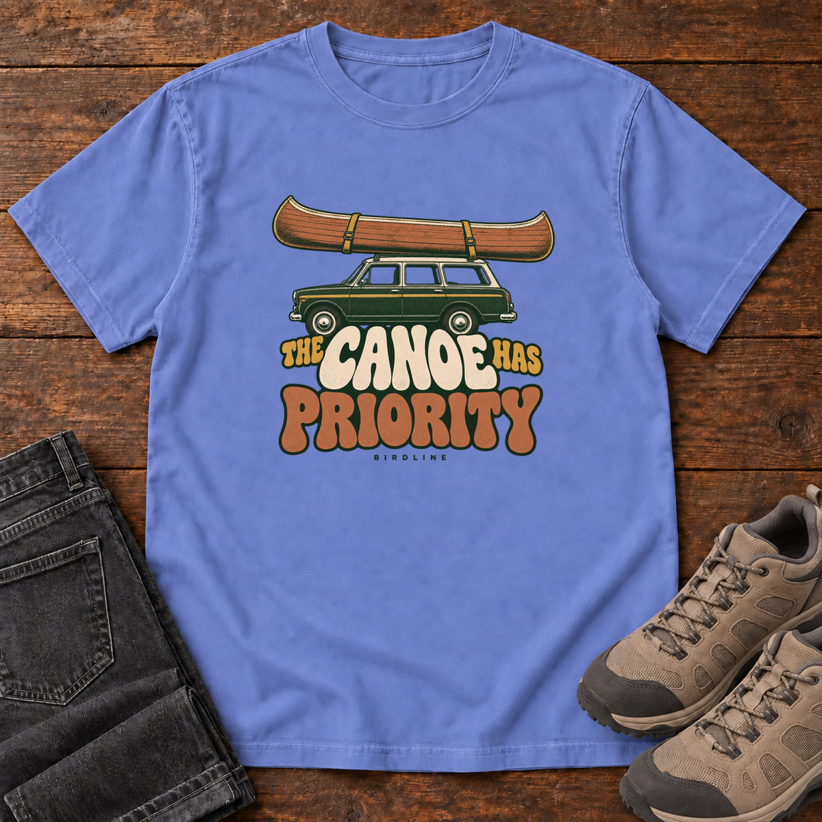 Flo Blue The Canoe Has Priority T-Shirt with a wooden canoe strapped atop a green station wagon over retro lettering; front flat lay on wood.