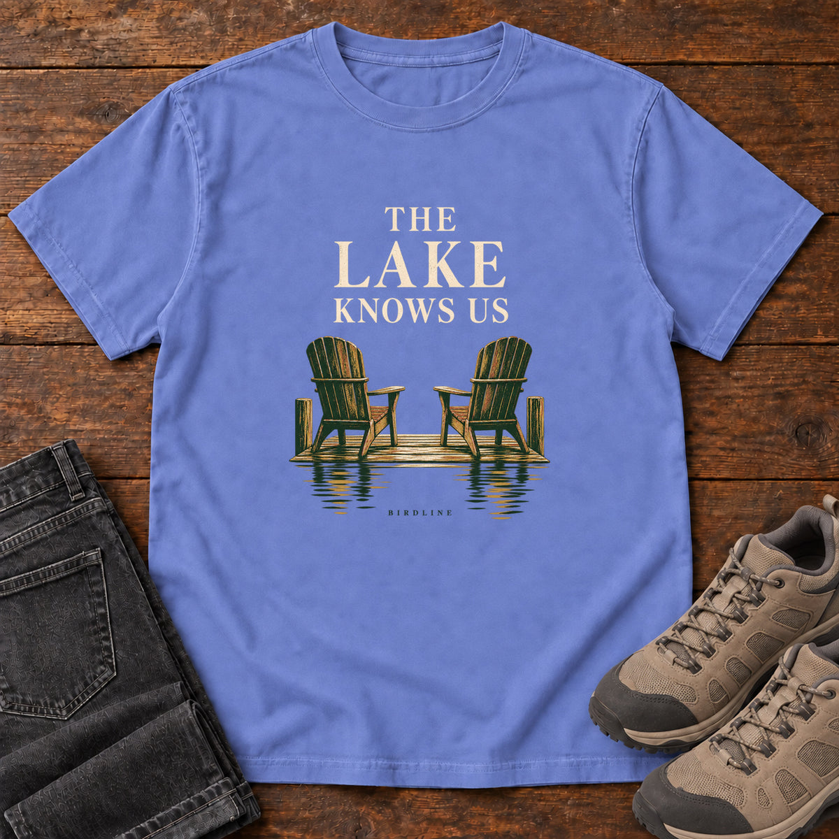 Flo Blue The Lake Knows Us T-Shirt with two green Adirondack chairs on a lakeside dock beneath cream lettering, front flat lay on wood.