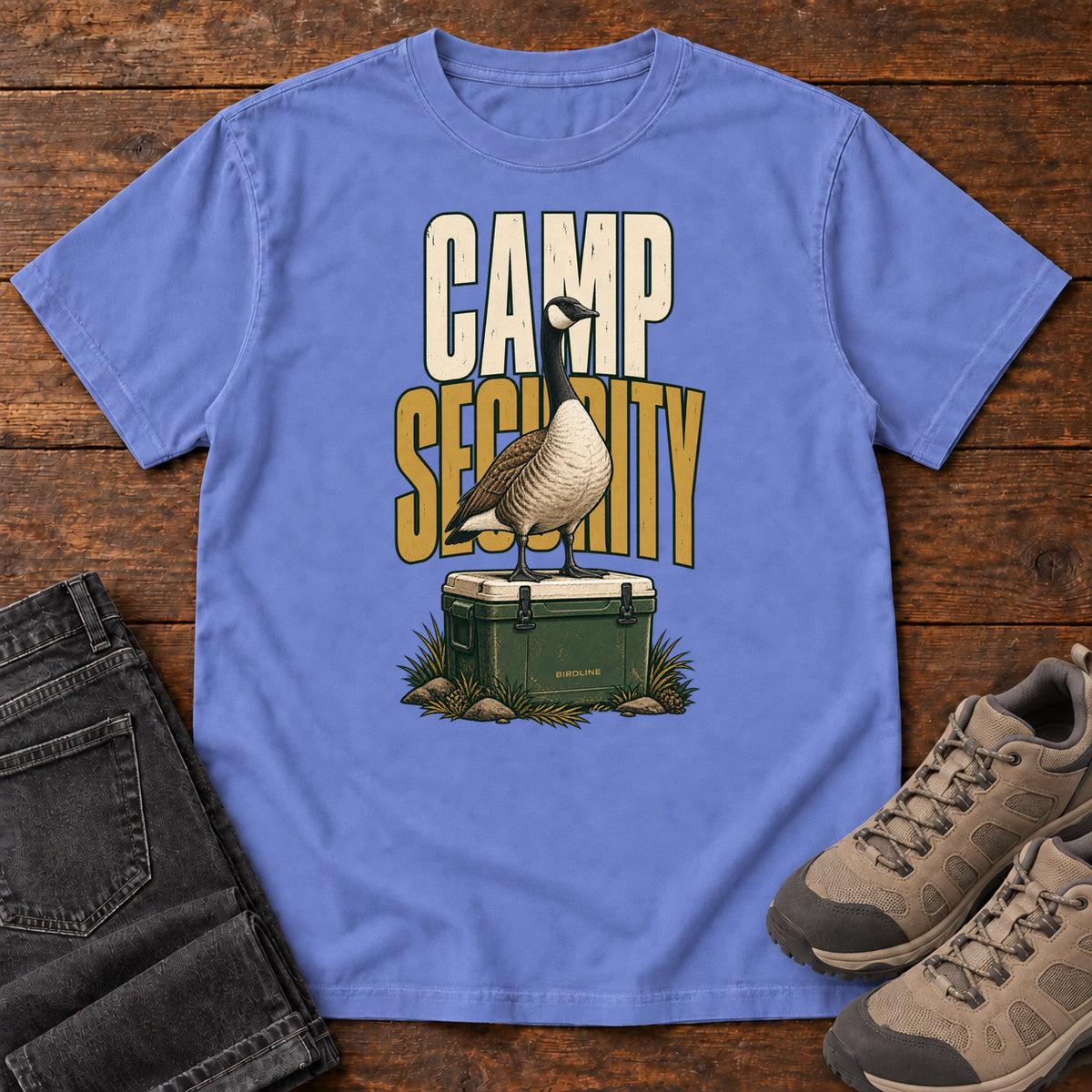 Flo Blue Camp Security T-Shirt with a goose standing on a green cooler beneath cream-and-gold lettering, front flat lay on wood.