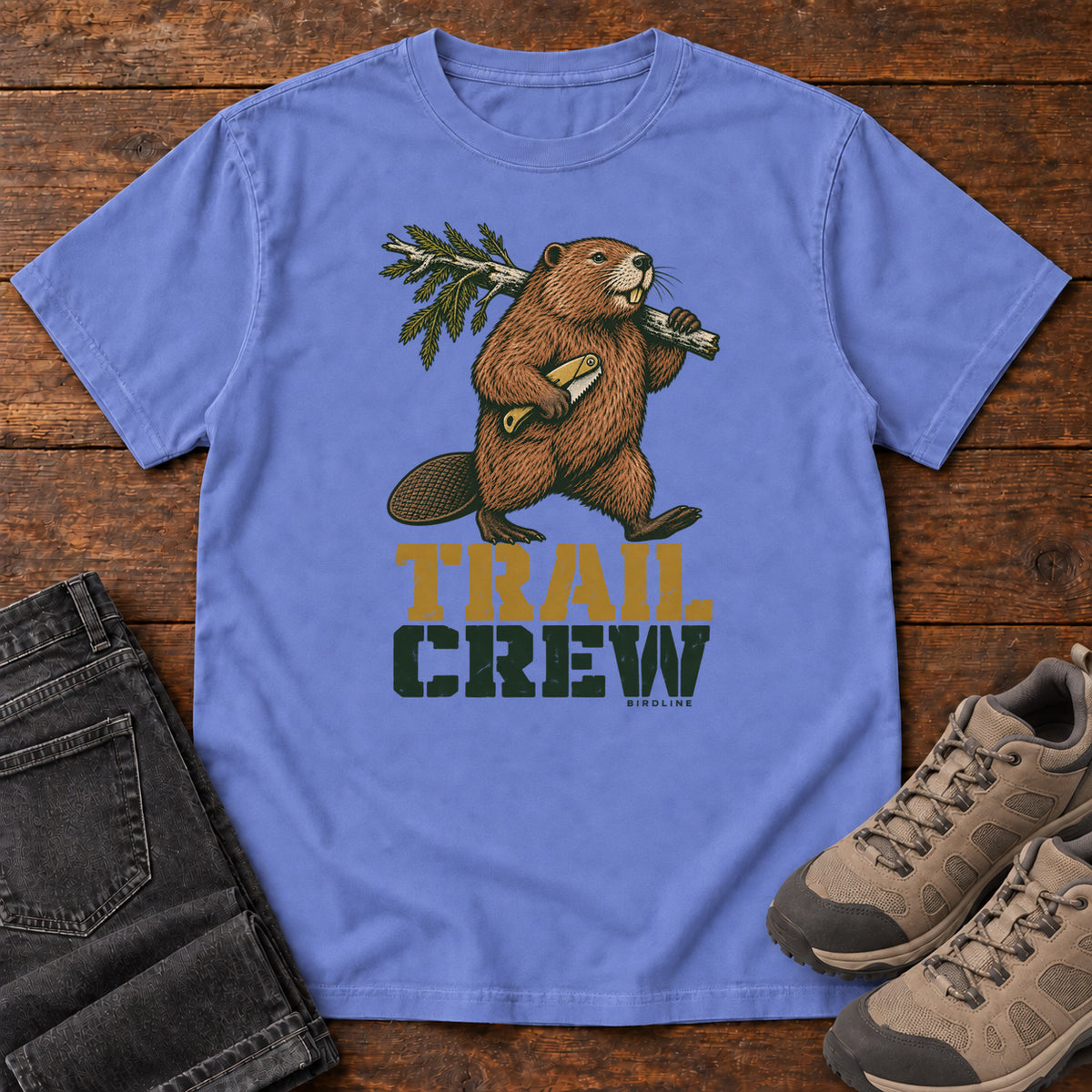 Flo Blue Trail Crew T-Shirt with a beaver carrying a leafy branch above gold and green Trail Crew lettering; front flat lay on wood.