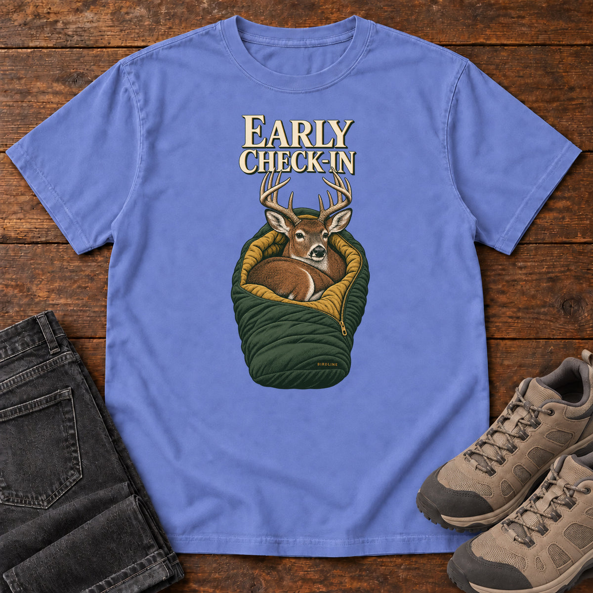 Flo Blue Early Check-In T-Shirt with an antlered deer curled inside a green sleeping bag; front flat lay on wood.