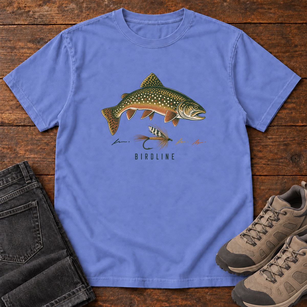 Flo Blue Trout and Fly T-Shirt with a green spotted fish above a fishing fly and Birdline lettering, front flat lay on wood.