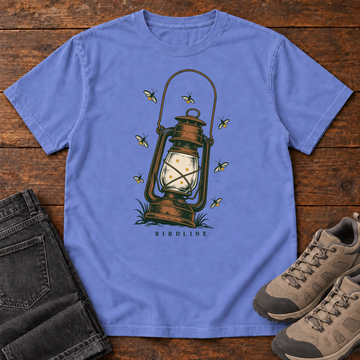 Flo Blue Lantern and Fireflies T-Shirt with a glowing vintage lantern surrounded by small flying insects; front flat lay on wood.