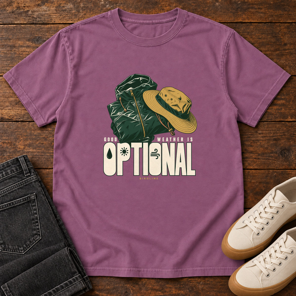 Good Weather Is Optional T-Shirt in Berry, front laid flat on wood, featuring a green hiking backpack and yellow brimmed hat above cream slogan lettering.