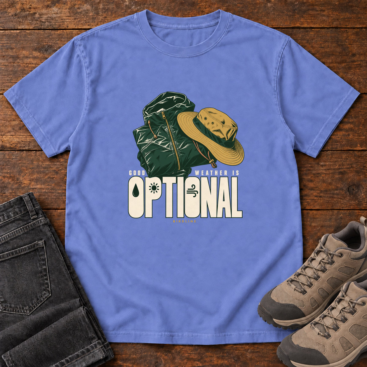 Good Weather Is Optional T-Shirt in Flo Blue, front laid flat on wood, featuring a green hiking backpack and yellow brimmed hat above cream slogan lettering.