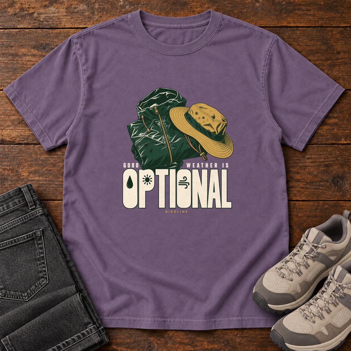 Good Weather Is Optional T-Shirt in Grape, front laid flat on wood, featuring a green hiking backpack and yellow brimmed hat above cream slogan lettering.