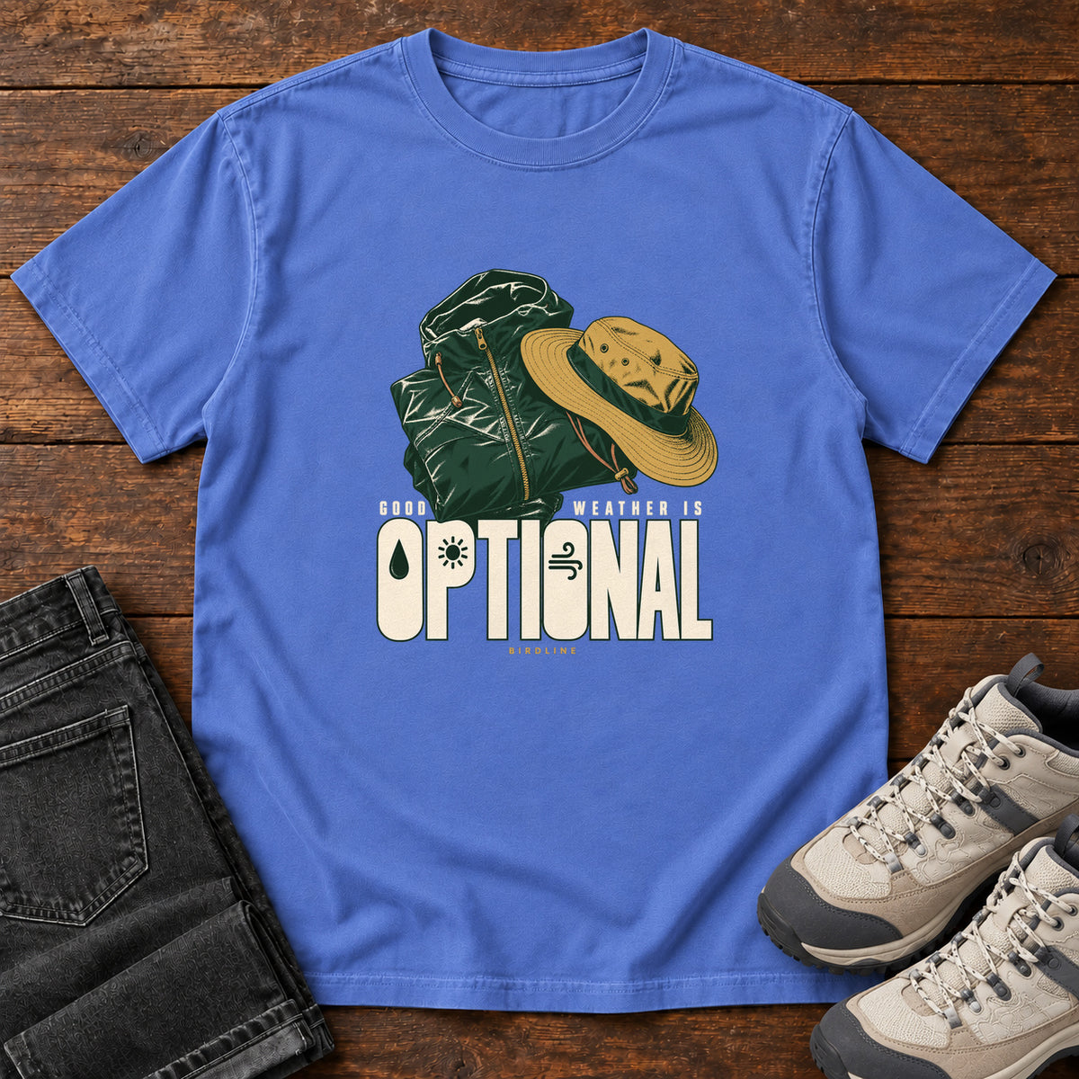 Good Weather Is Optional T-Shirt in Mystic Blue, front laid flat on wood, featuring a green hiking backpack and yellow brimmed hat above cream slogan lettering.