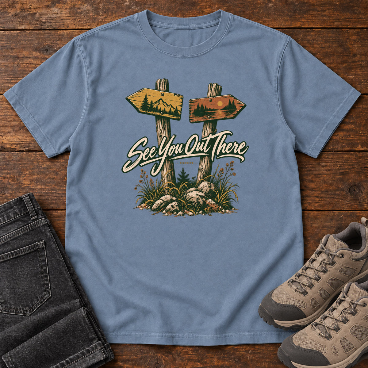Blue Jean See You Out There T-Shirt with two illustrated trail signposts, rocks, grass and cream script, front flat lay on wood.