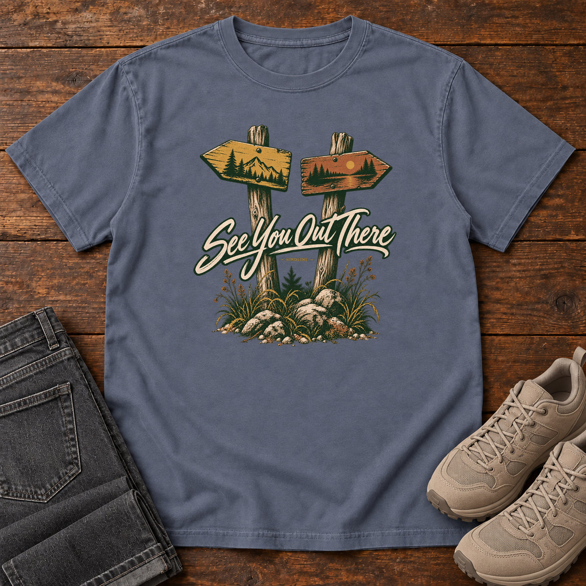 Denim See You Out There T-Shirt with two illustrated trail signposts, rocks, grass and cream script, front flat lay on wood.