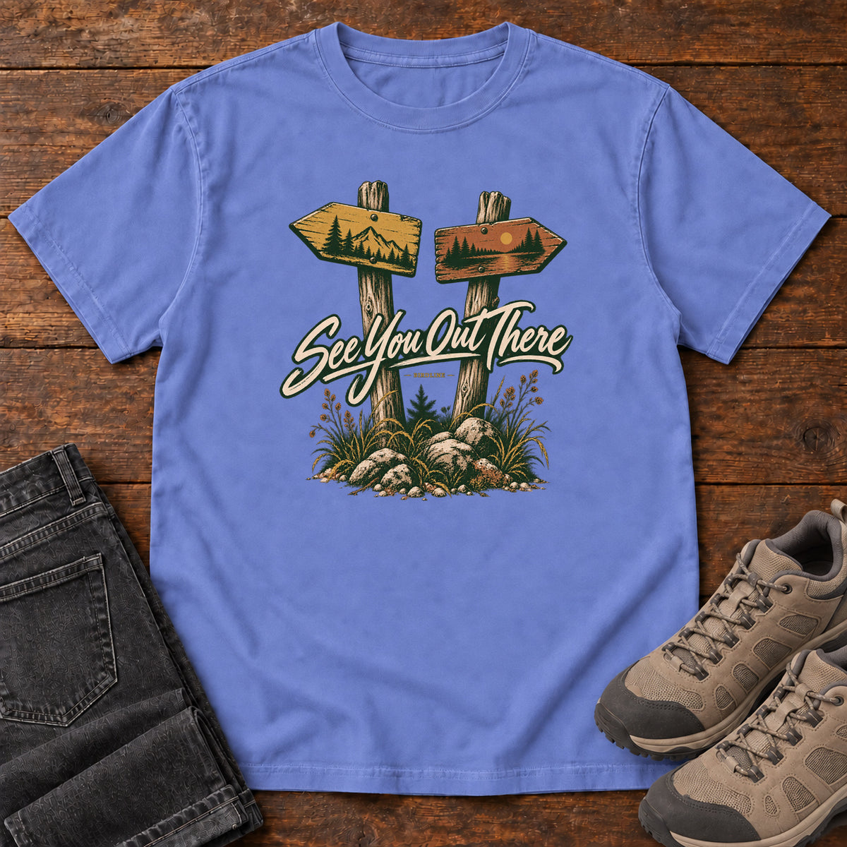 Flo Blue See You Out There T-Shirt with two illustrated trail signposts, rocks, grass and cream script, front flat lay on wood.